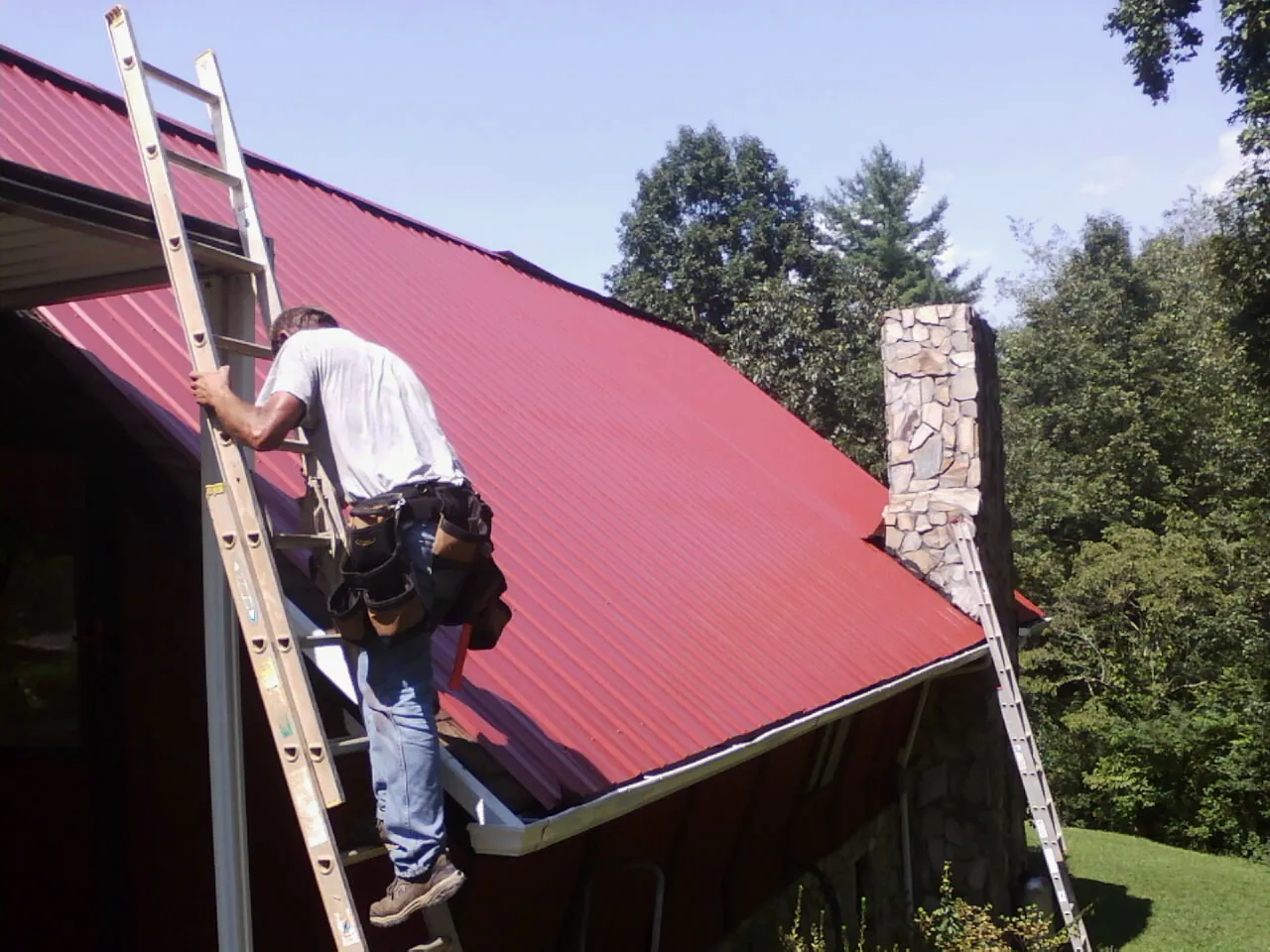 Roofer on a ladder beside a red metal roof and stone chimney — Soffit & Fascia Repair by Roofing Brothers in Pleasant Mount